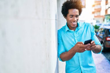 Genç, yakışıklı, Afro-Amerikan bir adam günlük giysiler giyip mutlu gülümsüyor. Yüzünde gülümsemeyle duvara yaslanıyorsun. Şehir caddesinde akıllı telefon kullanıyorsun..