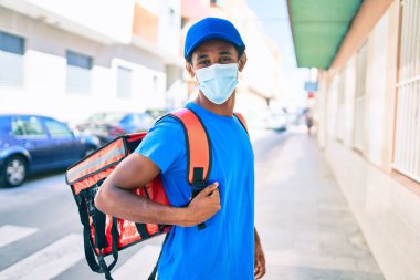Kurye üniforması giyen Afrikalı kurye sırt çantası takıyor. Koronavirüs güvenlik maskesi takıyor.