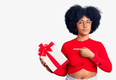 Genç Afrikalı Amerikalı kız elinde hediyeyle elini kenarı işaret ederek ciddi ve sakin bir yüz ifadesi sergiliyor. 