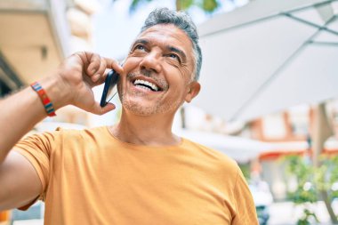 Orta yaşlı, kır saçlı, gülümseyen mutlu bir adam şehrin caddesinde akıllı telefondan konuşuyor..