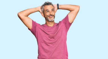 Orta yaşlı, gri saçlı, günlük kıyafetler giyen, rahatlayan ve esneyen, kolları ve elleri başının arkasında olan mutlu gülümseyen bir adam. 