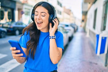 Genç İspanyol kadın şehirde akıllı telefon ve kulaklık kullanarak mutlu bir şekilde gülümsüyor..