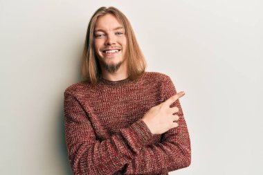 Uzun saçlı yakışıklı beyaz adam, günlük kışlık süveter giyiyor. Gülümsüyor, eli ve parmağı yana doğru uzanıyor. 