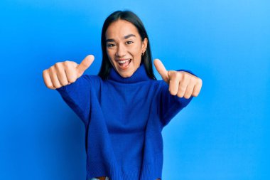 Asyalı genç bir kadın günlük kışlık süveter giyiyor. Elleriyle olumlu bir jest yapıyor. Gülümsüyor ve başarı için seviniyor. kazanan jest. 