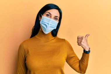 Tıbbi maske takan genç beyaz bir kadın baş parmağını yukarı kaldırıyor ve ağzı açık gülümsüyor. 