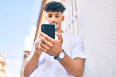 Genç Arap adam mutlu bir şekilde gülümsüyor. Duvara yaslanmış akıllı telefon kullanıyor..