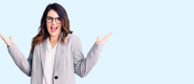 İş kıyafetleri ve gözlük takan güzel esmer kadın zaferi mutlu bir gülümsemeyle kutluyor ve ellerini kaldırarak kazanan yüz ifadesi takınıyor. 