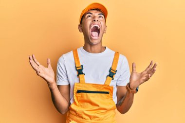 Sarı arka plan üzerine tamirci üniforması giyen yakışıklı Afro-Amerikalı genç adam deli gibi bağırıyor ve agresif bir ifadeyle bağırıyor ve kollarını kaldırıyor. hüsran kavramı. 