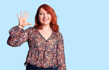 Genç, güzel, kızıl saçlı, gündelik tişört giyen, beş numaralı parmaklarıyla işaret eden, kendinden emin ve mutlu bir şekilde gülümseyen kadın.. 