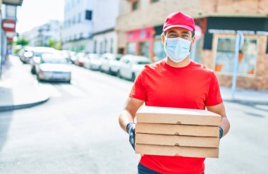 Üniforma giyen ve koronavirüs koruma maskesi takan genç teslimatçı. Kasaba caddesinde pizza kutularını teslim ediyorum..