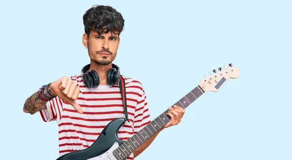 怒った顔でエレキギターを弾く若いヒスパニック系の男 親指で嫌いを示す負の記号 拒否の概念 — ストック写真