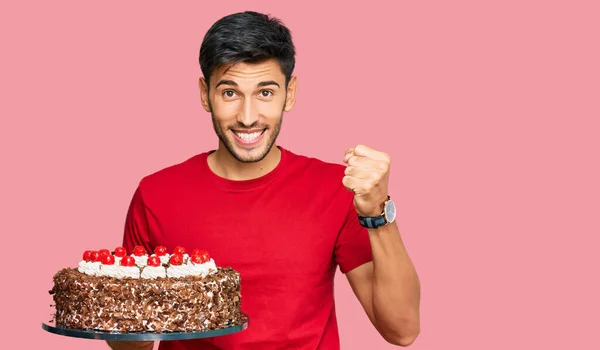 Young handsome man celebrating birthday with cake screaming proud, celebrating victory and success very excited with raised arms