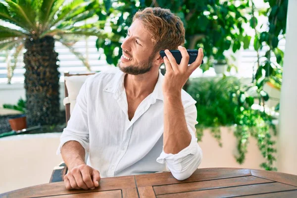 Yakışıklı beyaz adam terasta oturmuş gülümsüyor ve telefondaki bir sesli mesajı dinliyor.
