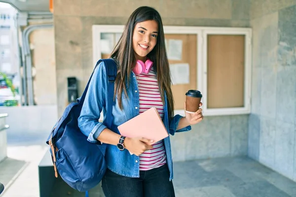 Young hispanic student girl smiling happy holding book and drinking coffee at the university.