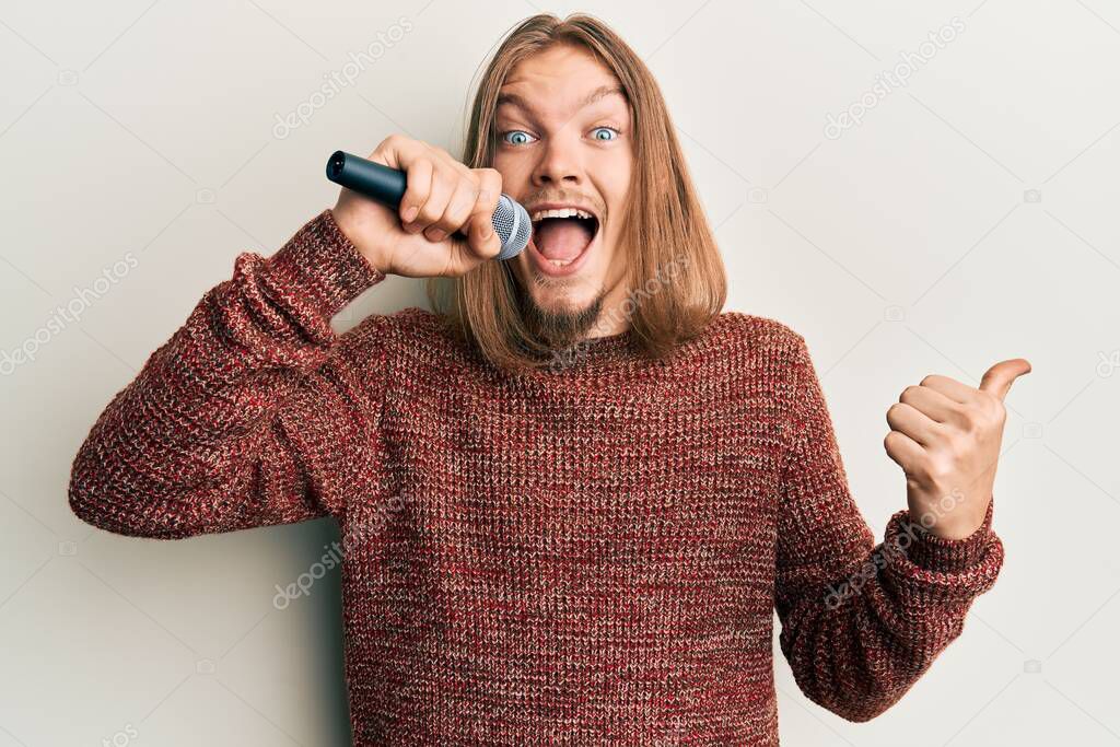 Handsome caucasian man with long hair singing song using microphone pointing thumb up to the side smiling happy with open mouth 