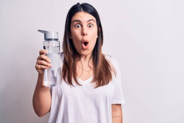 Genç, güzel esmer kadın izole edilmiş beyaz arka planda bir şişe su içiyor. Şaşırmak için ağzı açık ve korkmuş, inançsız bir yüz.
