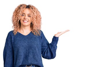 Kıvırcık saçlı, sıradan bir kazak giyen neşeli bir şekilde gülümseyen ve avuç içi kameraya bakan genç sarışın kadın.. 