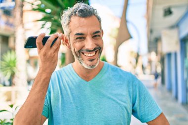 Orta yaşlı, kır saçlı, akıllı telefondan konuşan bir adam şehrin caddesinde yürüyor..
