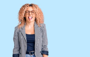 Kıvırcık saçlı, iş ceketi giyen ve gözlüklü, dilini dışarı çıkaran neşeli ifadeli genç sarışın kadın. duygu kavramı. 