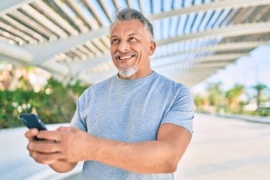 Orta yaşlı, ispanyol, kır saçlı, mutlu gülümseyen bir adam parkta akıllı telefon kullanıyor..