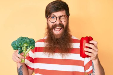 Uzun sakallı, elinde brokoli ve kırmızı biber olan yakışıklı genç adam kameraya seksi bir ifadeyle bakıyor, neşeli ve mutlu bir yüz.. 