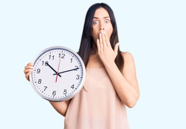 Genç, beyaz bir kadın büyük bir saat tutuyor. Ağzını eliyle kapatıyor. Şok olmuş ve hata yapmaktan korkuyor. şaşırmış ifade 