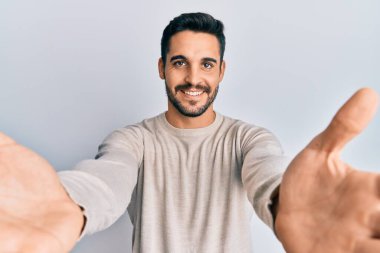 Gündelik giysiler giymiş genç İspanyol adam kameraya bakıp sarılmak için kollarını açmış gülümsüyor. Mutluluğu kucaklayan neşeli bir ifade.. 