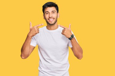 Gündelik beyaz tişört giyen, neşeli bir şekilde gülümseyen ve parmaklarıyla ve ağzıyla işaret eden genç yakışıklı bir adam. diş sağlığı kavramı. 