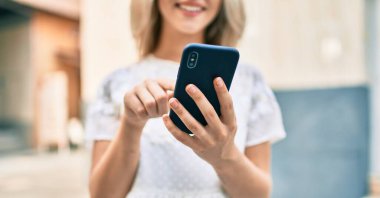 Beyaz kız şehrin caddesinde akıllı telefon kullanarak mutlu bir şekilde gülümsüyor..