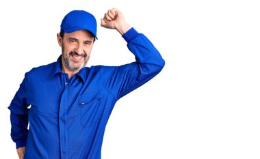 Orta yaşlı yakışıklı, tamirci üniforması giyen, gülümseyen, kendine güvenen, el hareketleriyle saçlara dokunan çekici ve modaya uygun poz veren bir adam. 