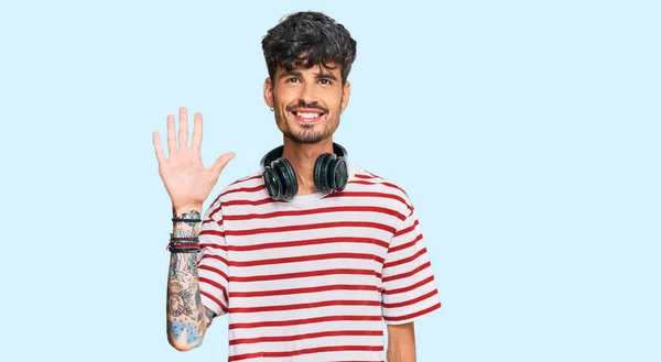 自信と幸せな笑顔ながら 若いヒスパニック系の男は ヘッドフォンを使用して音楽を聴くと指の数を示すと指摘します — ストック写真