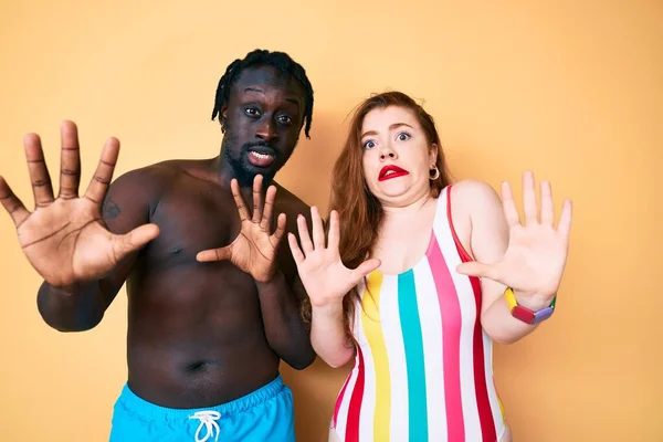 Interracial couple wearing swimwear afraid and terrified with fear expression stop gesture with hands, shouting in shock. panic concept.