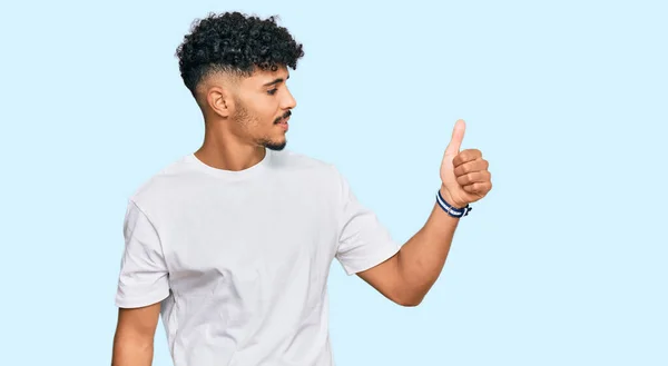 若いアラブ人男を着てカジュアルな白いTシャツを誇りに思って見て 笑顔をやって親指を横にジェスチャー — ストック写真