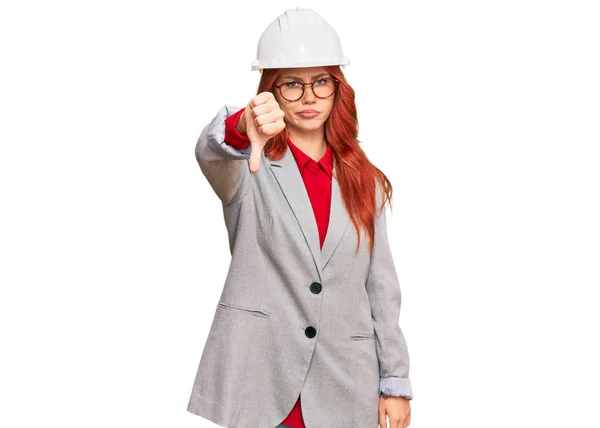 年轻的红头发女人戴着建筑师的硬礼帽 看上去很不高兴 很生气 用大拇指垂下的姿势表示拒绝和消极 不好的表达方式 — 图库照片