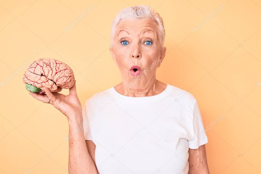 Senior beautiful woman with blue eyes and grey hair holding brain as mental health concept scared and amazed with open mouth for surprise, disbelief face 