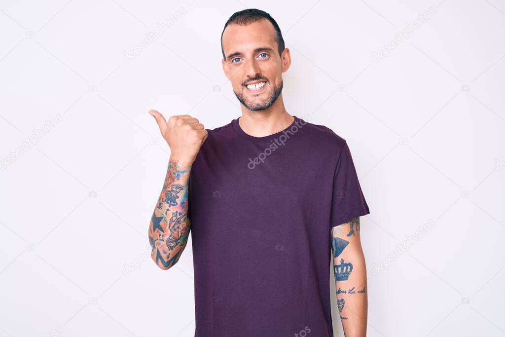 Young handsome man with tattoo wearing casual clothes smiling with happy face looking and pointing to the side with thumb up. 