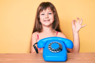 Masada oturan uzun saçlı beyaz bir kız antika telefon kullanıyor. Parmaklarıyla işaret veriyor, gülümsüyor. 