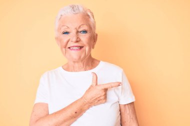 Son sınıf güzeli, mavi gözlü, gri saçlı, sarı arka planda klasik beyaz tişörtlü, neşeli, yüzünde mutlu ve doğal bir ifade olan el ve parmak ile yüzünü gösteren bir gülümsemesi olan bir kadın. 