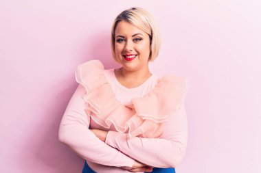 Genç, güzel, sarışın ve cüsseli bir kadın izole edilmiş pembe arka plan üzerine sıradan bir kazak giyip kollarını kavuşturup kameraya bakıyor. Pozitif kişi.