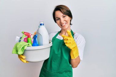 Kısa saçlı, esmer, önlüklü, temizlik malzemesi tutan, mutlu ve komik yüzlü genç bir kadın. iyi enerji ve titreşimler. 