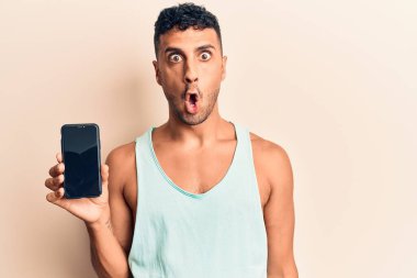 Akıllı telefonu tutan genç İspanyol adam ekranda korkmuş ve ağzı açık bir şekilde şaşırmış, inançsız bir yüz ifadesi sergiliyor. 