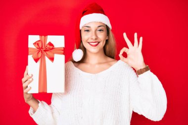 Genç esmer kadın noel şapkası takıyor ve elinde bir hediye tutuyor tamam işareti yapıyor parmaklarıyla, gülümseyerek mükemmel bir sembol oluşturuyor 