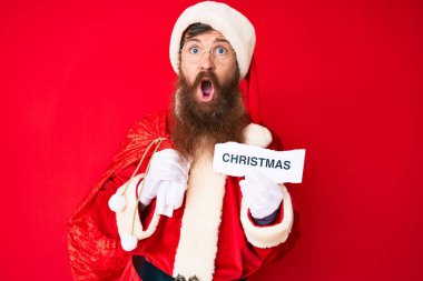 Uzun sakallı yakışıklı kızıl saçlı adam Noel Baba kostümü giyiyor. Elinde yılbaşı mesajı var. Korkmuş ve şok olmuş bir halde.. 