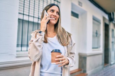 Beyaz kadın gülümsüyor. Akıllı telefondan konuşuyor ve şehirde kahve içiyor..