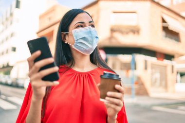 Tıbbi maske takan genç Latin kız akıllı telefon kullanıyor ve şehirde kahve tutuyor..