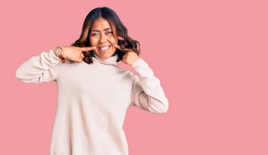 Balıkçı yaka kazak giyen, neşeli gülümseyen ve parmakları, dişleri ve ağzıyla işaret eden genç ve güzel bir ırk kadını. diş sağlığı kavramı. 