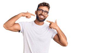 Gündelik kıyafetler giyen ve gözlüklü, neşeli gülümseyen, parmaklarıyla ve ağzıyla işaret eden İspanyol genç bir adam. diş sağlığı kavramı. 