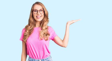 Sarı saçlı, sıradan giyinmiş, gözlüklü, neşeli gülümseyen, eli kameraya bakan güzel beyaz bir kadın.. 