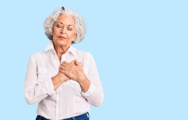 Üstün gri saçlı, günlük kıyafetler giyen, elleri göğsünde, gözleri kapalı gülümseyen ve yüzünde minnet ifadesi olan bir kadın. sağlık kavramı. 