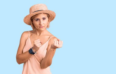 Sarışın, yaz şapkası takan genç bir kadın. Yumruk savunma hareketiyle dövüşmeye hazır. Kızgın ve üzgün bir yüz. 